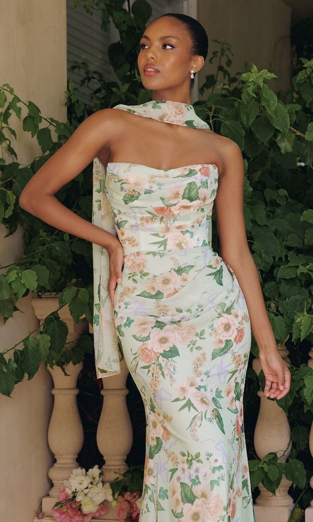 A woman stands before green foliage wearing the Amoris T718 Strapless Print Mermaid Prom Gown with a floral pattern and a matching scarf around her neck.